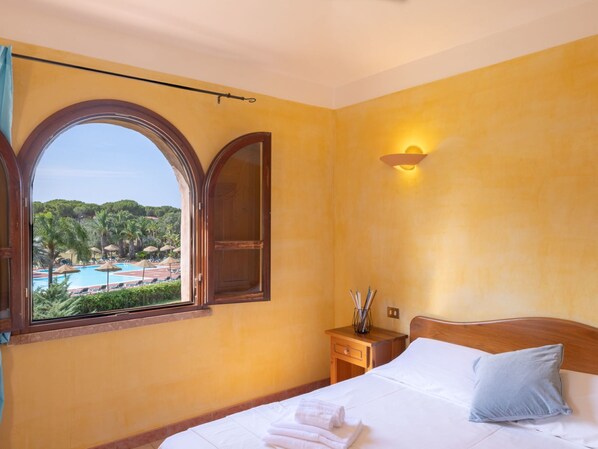 A hotel room with a large window overlooking a pool and greenery, a bed with white linens, and a nightstand with a lamp.