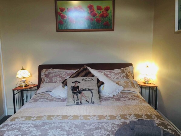 Una cama bien hecha con ropa de cama floral, dos lámparas de noche y un cuadro con flores rojas encima.