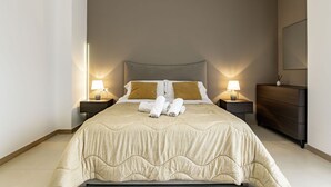 Quarto de hotel com uma cama grande, duas mesas de cabeceira com luminárias, um aparador de madeira escura e um espelho grande na parede.