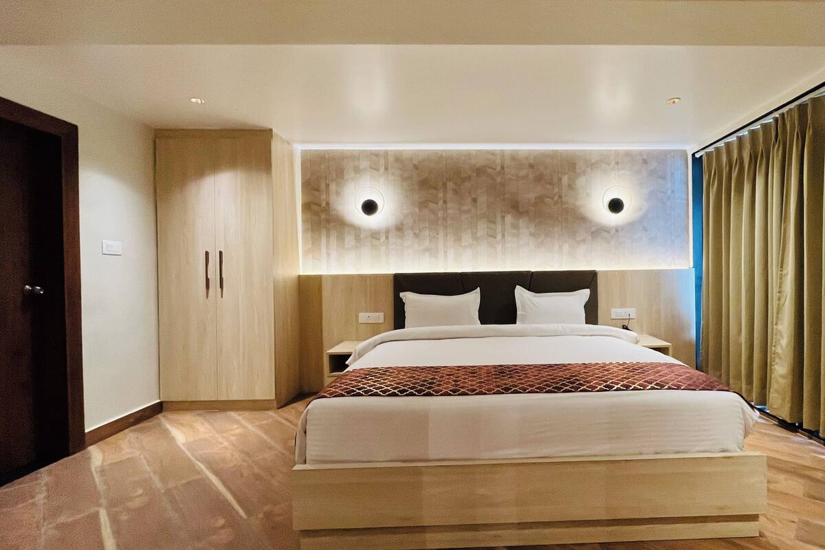 A modern hotel room with a large bed, wooden flooring, and a view of the city through floor-to-ceiling windows.