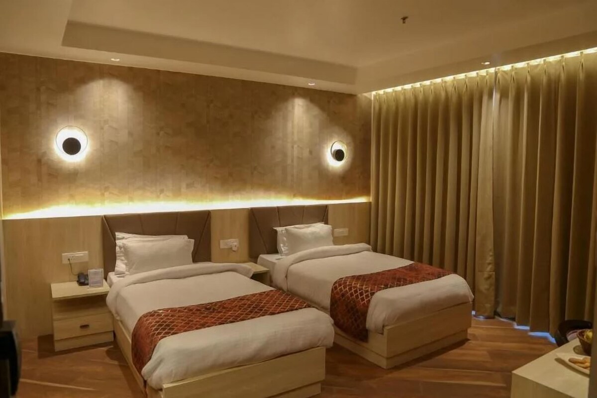 A hotel room with two beds, a large wall-mounted light fixture, a window with curtains, and a wooden floor.