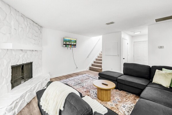 Una sala de estar moderna con chimenea, un sofá negro, un tapete estampado y una escalera que conduce a otra habitación.