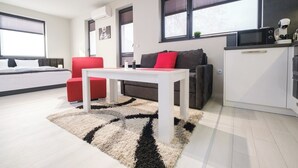 Een moderne woonkamer met een witte tafel, een rood-grijze bank en een keukenhoek met een magnetron en waterkoker.