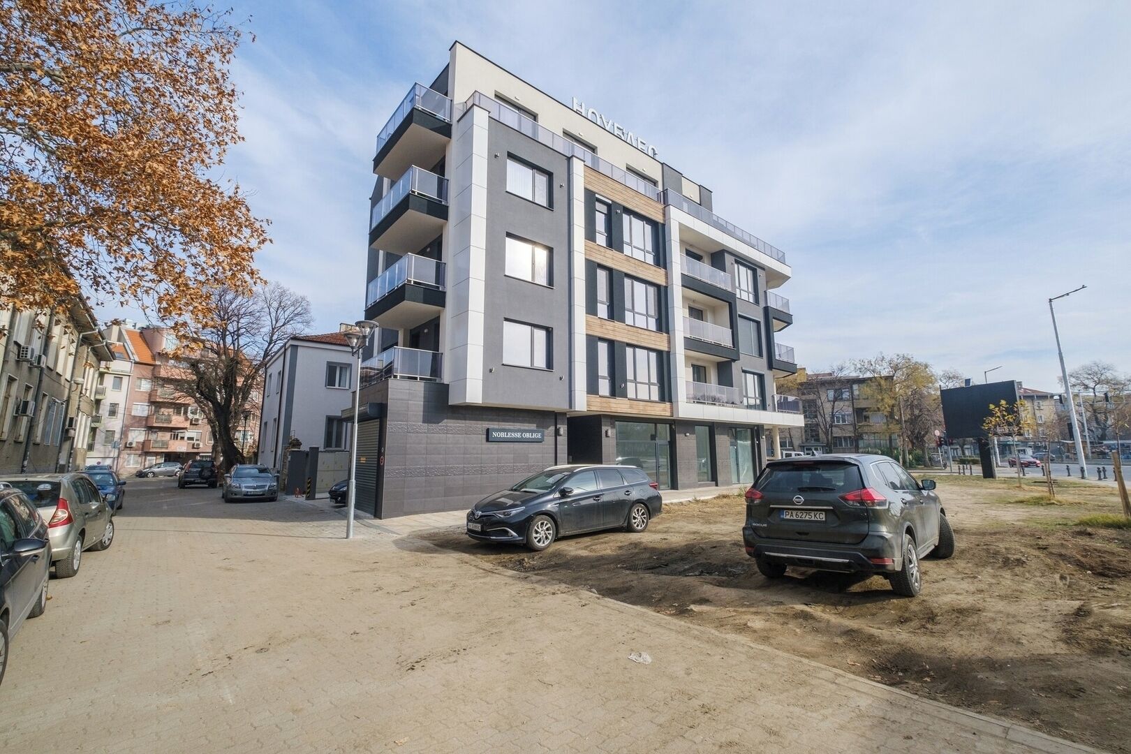 Un edificio residencial de varios pisos con balcones, un área de estacionamiento y un sendero de tierra.