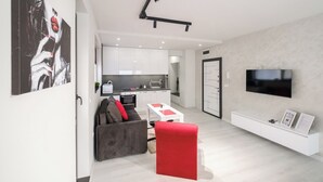 Een moderne woonkamer met een grijze bank, een rode ottoman en een witte salontafel. Een flatscreen televisie is aan de muur bevestigd, en een keuken is op de achtergrond zichtbaar.