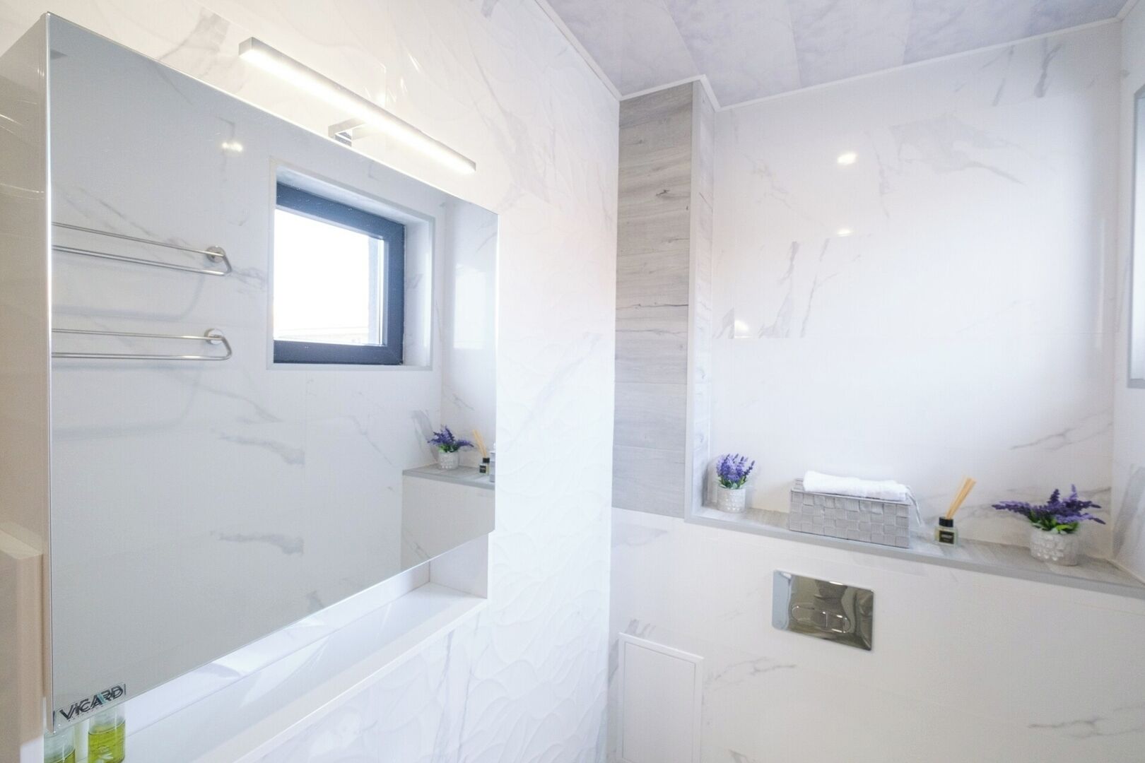 Un baño moderno con un espejo grande, una ventana y un estante empotrado con artículos de aseo.
