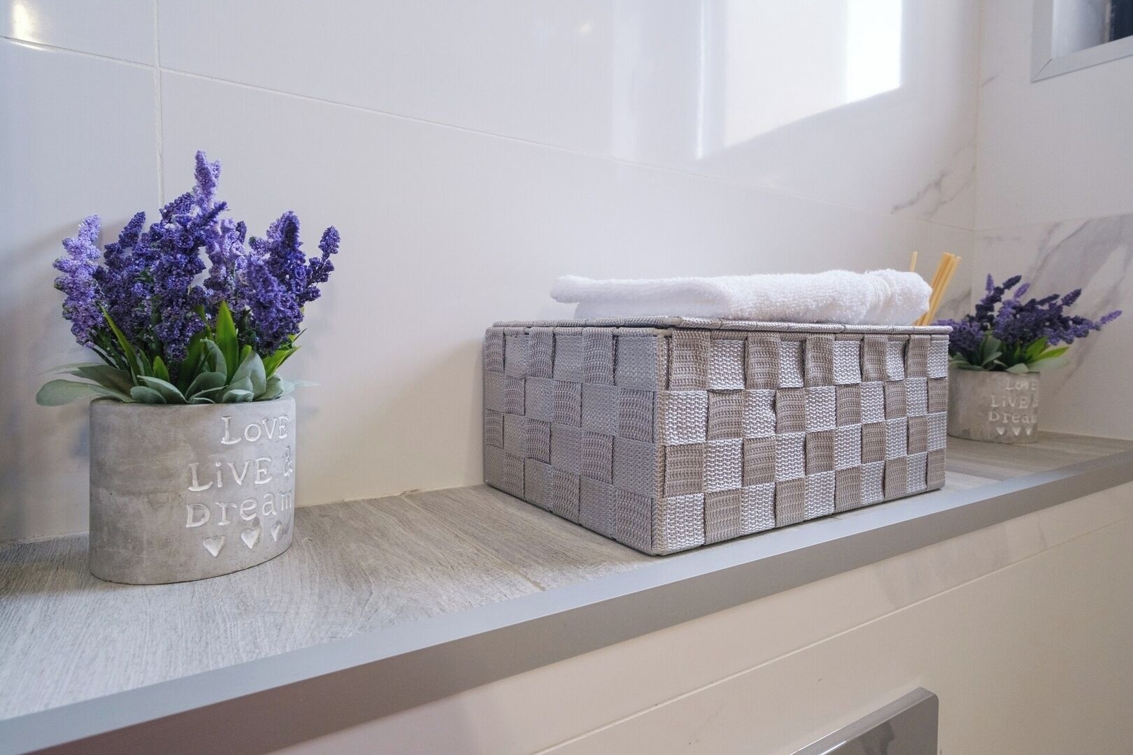 Un baño con pared de azulejos blancos, una cesta trenzada gris en un estante y dos macetas con plantas de lavanda.