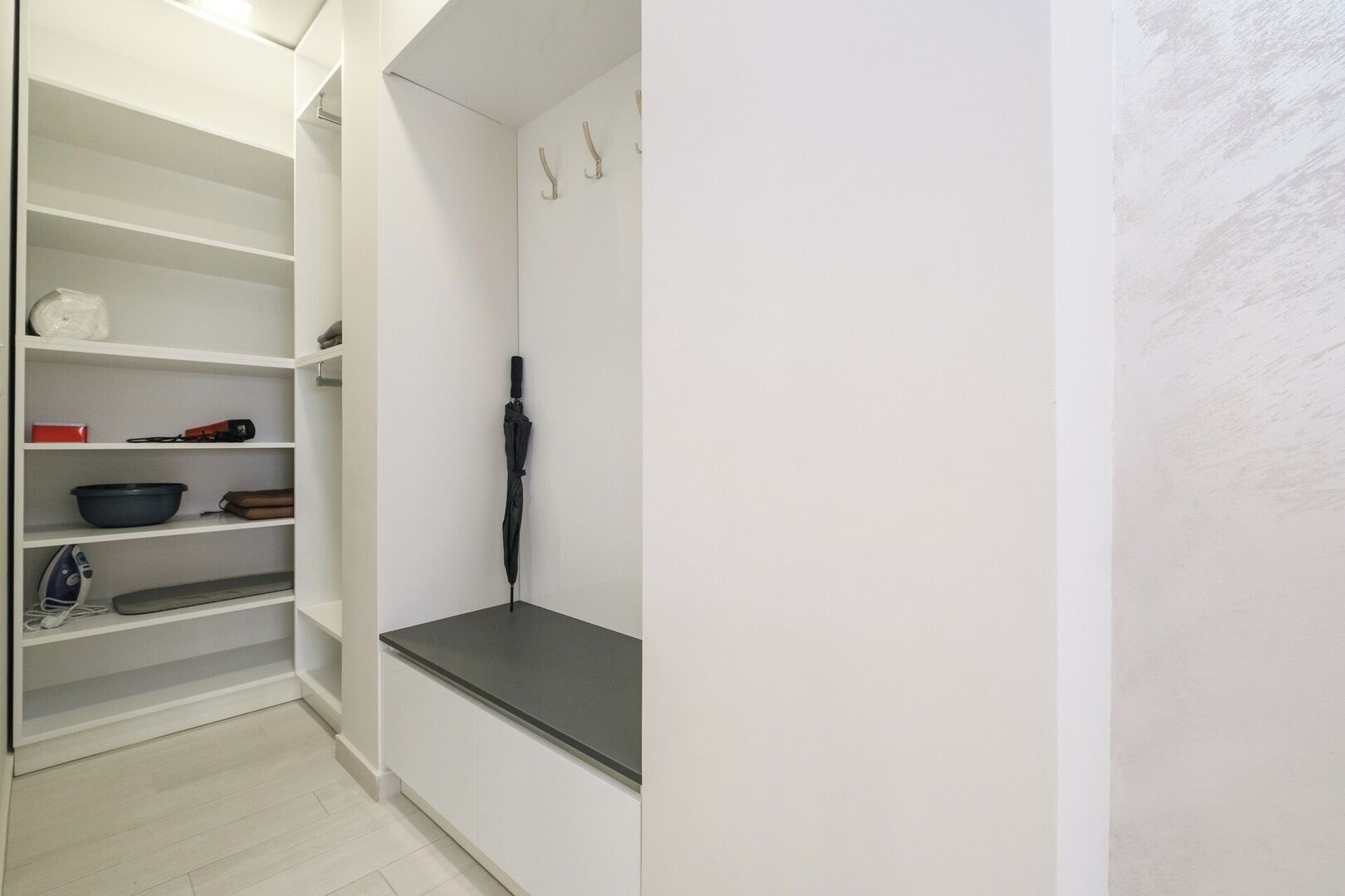 Ein weißer Schrank mit Regalen, in denen sich eine rote Kiste, eine schwarze Schüssel und ein Bügeleisen befinden.