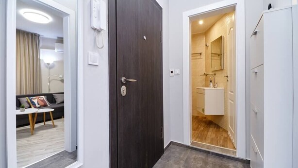 A hotel room with a dark wooden door, a bathroom with a white sink and mirror, and a living area with a sofa and a small table. - Cozy 1-bed Apartment 2 (Plovdiv)