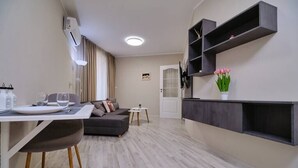 A modern living room with a grey sofa, a dining table with chairs, a small white table, and a wall-mounted shelf with a vase of flowers. - Cozy 1-bed Apartment 2 (Plovdiv)