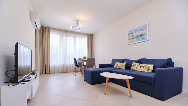 A modern living room with a blue sofa, a small white table, a TV, and a dining area with chairs and a table. - Cozy 1-bed Apartment (Plovdiv)