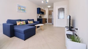 A modern living room with a blue sofa and a small white table. A kitchen is visible in the background with wooden cabinets and a refrigerator. - Cozy 1-bed Apartment (Plovdiv)