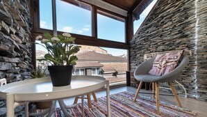 Ein Zimmer mit Steinsandsteinwand, einem großen Fenster, einem runden Tisch mit Topfpflanze, einem grauen Sessel mit gemustertem Kissen und einem farbenfrohen Streifen Teppich.