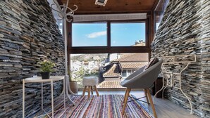 Ein gemütliches Zimmer mit Steinwänden, einem großen Fenster, einem Sessel, einem kleinen Tisch und einem farbenfrohen Teppich.