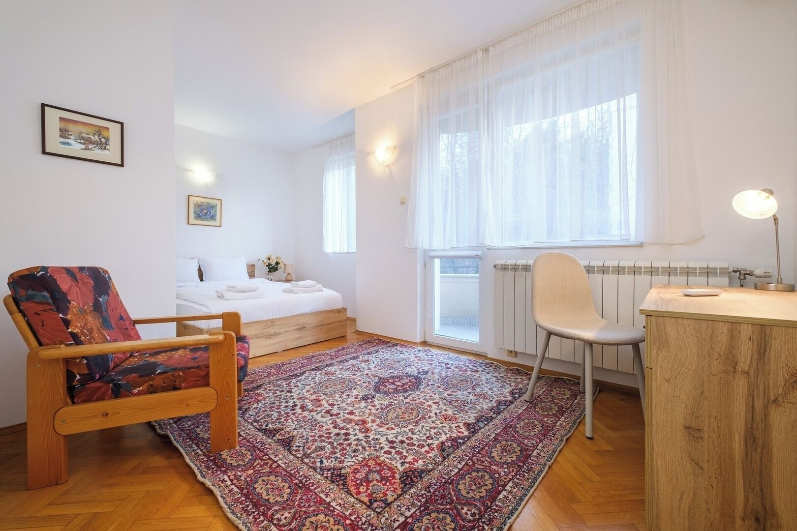 A bedroom with a bed, a chair, a desk, a radiator, and a window with sheer curtains.
