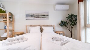 A hotel room with two beds, a wooden headboard, a painting on the wall, a plant, and a shelf with towels and a basket.