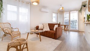 A spacious living room with a brown sofa, wicker chairs, a glass coffee table, and a large window providing natural light.