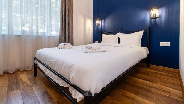 A neatly made bed with white linens and towels, a dark blue wall, and a wooden floor.