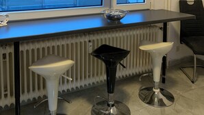 A modern kitchen with a black countertop, white bar stools, a glass bowl, and a window with blinds. - Accogliente appartamento (Wuppertal)