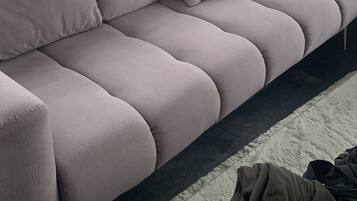 Ein graues Sofa mit vielen Kissen, eine TV-Konsole mit schwarzem Rahmen und ein weißer, flauschiger Teppich.