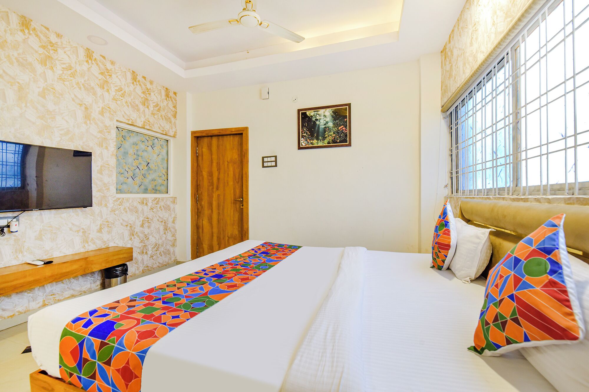 Habitación de hotel con cama, televisor, banco de madera, puerta y ventana con barrotes.