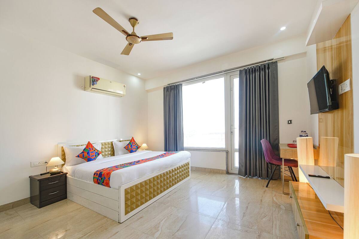 A hotel room with a bed, bedside table, lamp, air conditioning unit, ceiling fan, large window with curtains, and a TV mounted on the wall.