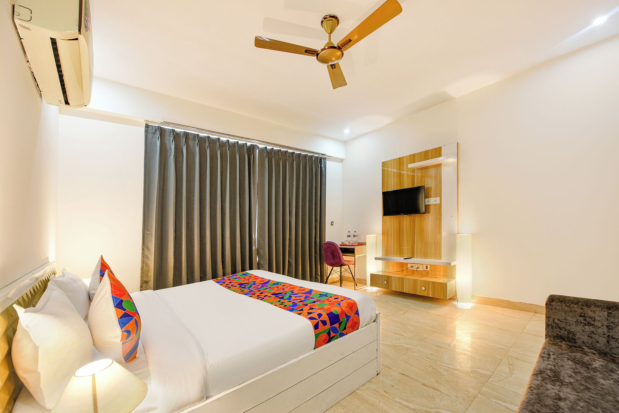 Ein Hotelzimmer mit Bett, Schreibtisch, Stuhl, Wandfernseher und Deckenventilator.