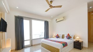 A hotel room with a bed, bedside tables, a ceiling fan, a window with curtains, and an air conditioning unit. - Fabhotel Ar Suites (Gurugram)