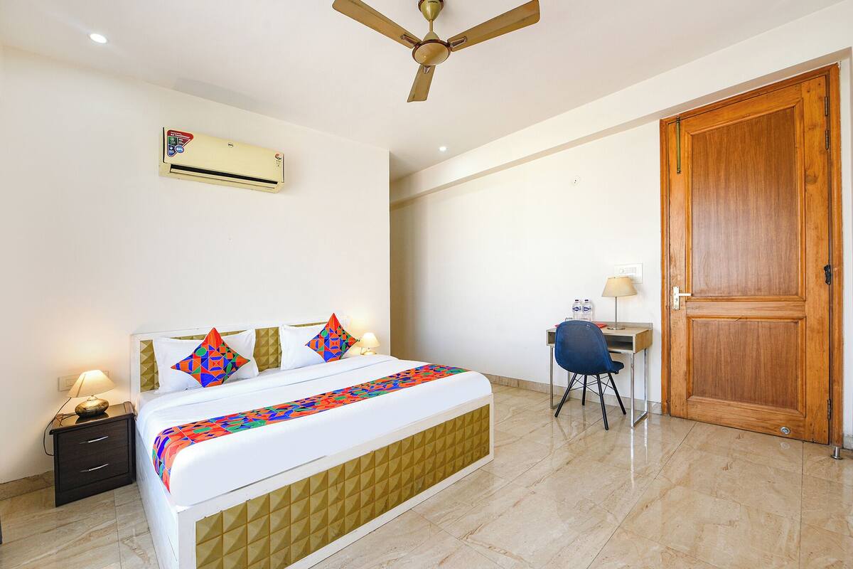 A hotel room with a bed, a desk, a chair, an air conditioner, and a wooden door.