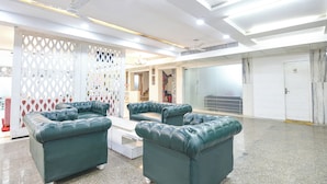 Een ruime lobby met een hoge plafond, een zithoek met groene gestoffeerde banken, een witte salontafel en een decoratieve witte spijlenwand.