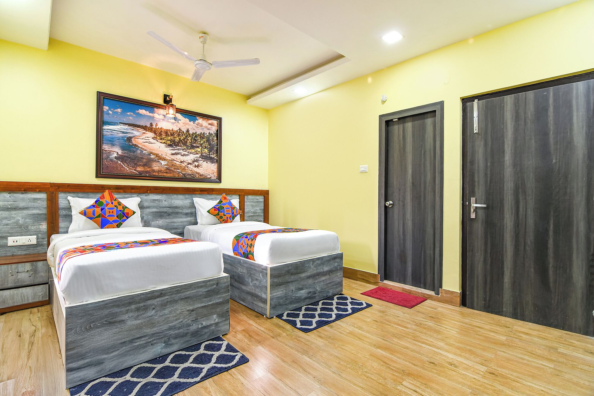 A hotel room with two beds, a ceiling fan, a painting, and two doors.