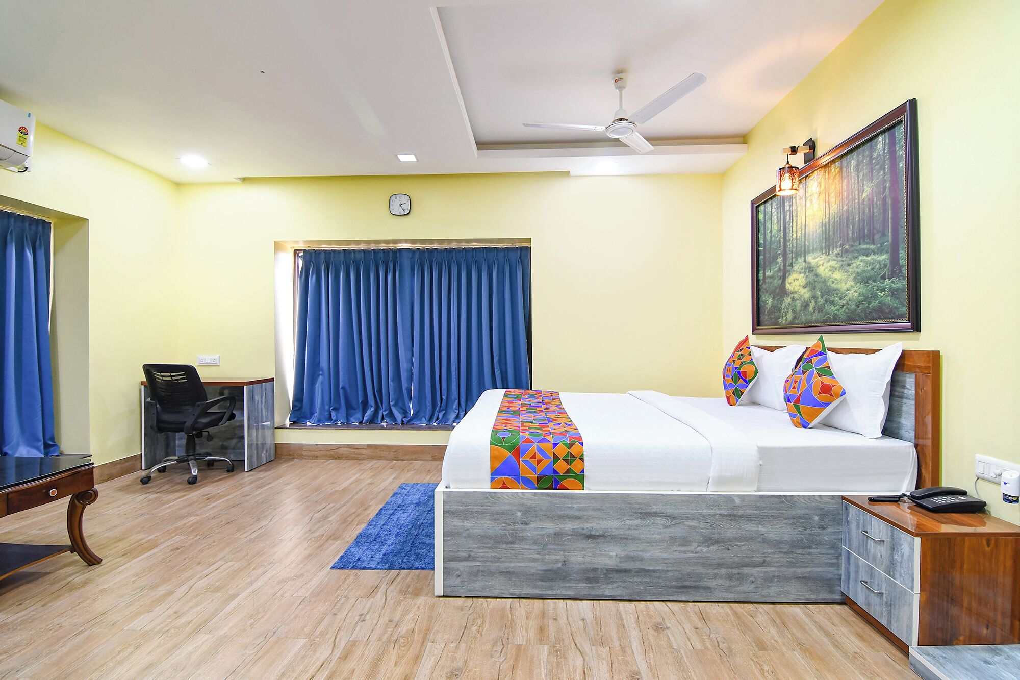 A bedroom with a bed, a desk, a chair, a ceiling fan, and a painting on the wall.