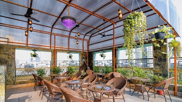 An outdoor seating area with wicker chairs and tables, surrounded by potted plants and a metal roof structure. - Fabhotel Prime Familto (Baruipur)