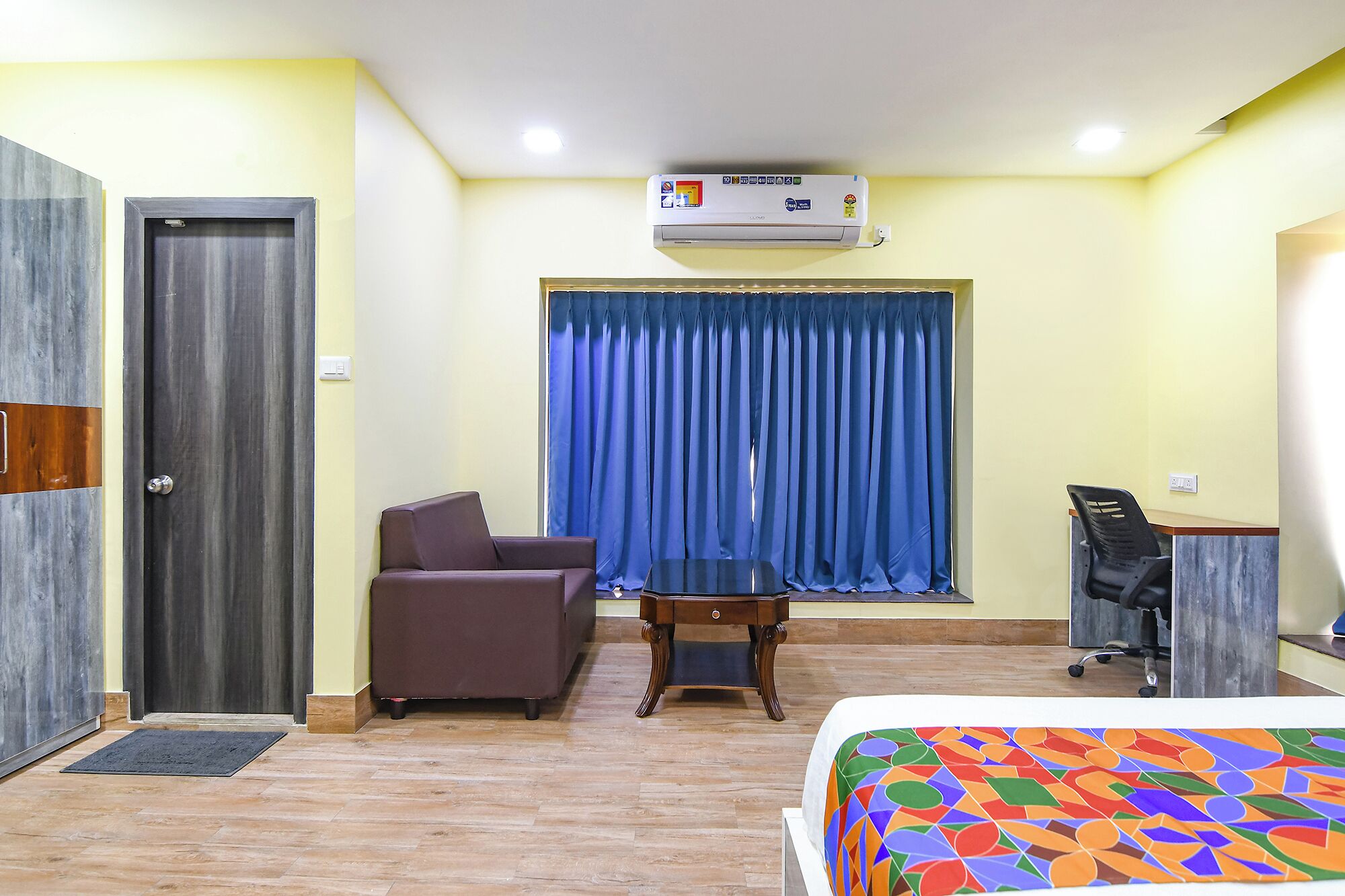 A hotel room with a bed, a desk, a chair, a sofa, a window with curtains, an air conditioning unit, and a door.