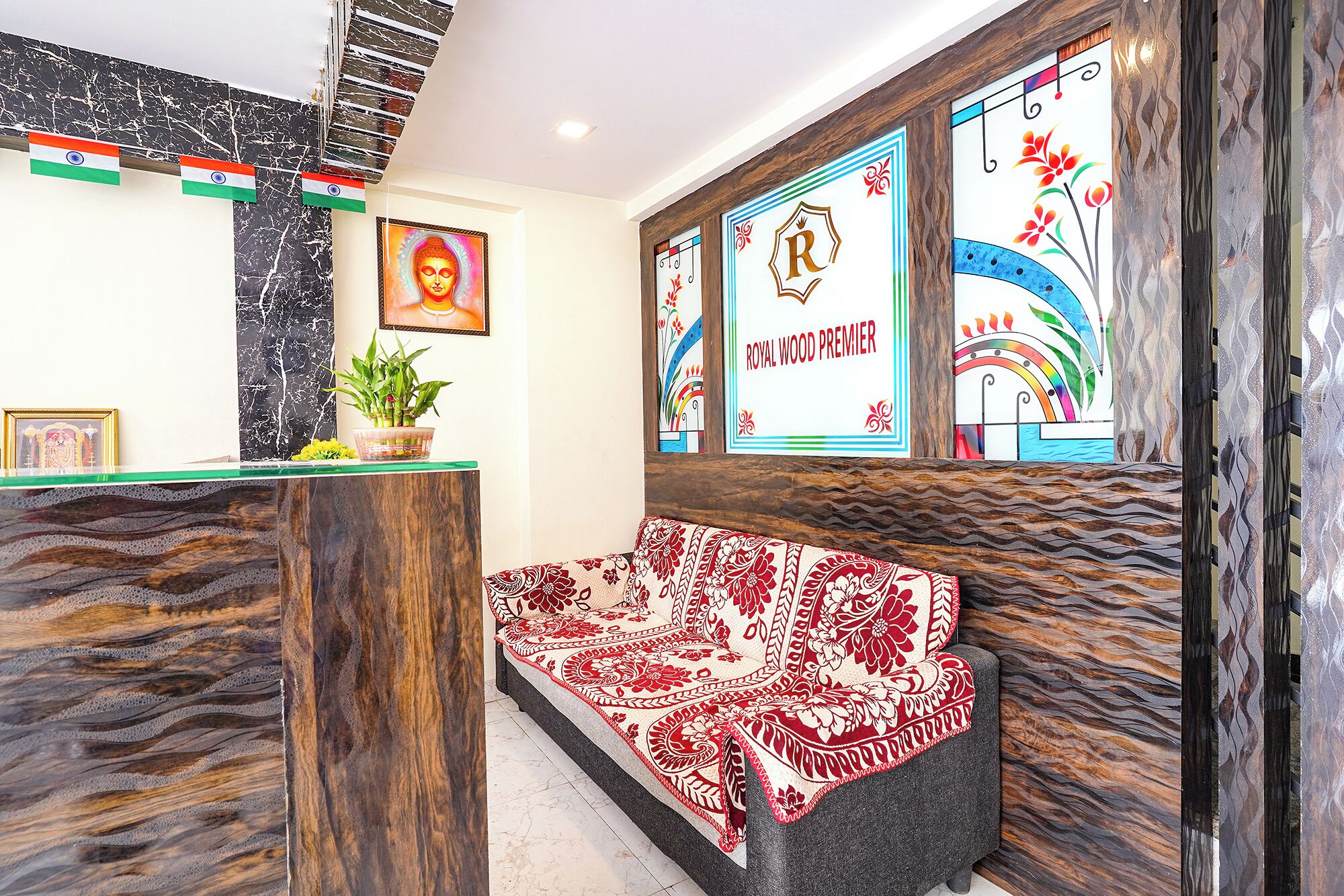 Un área de recepción con un mostrador de madera, un banco con cojín estampado en rojo y blanco, y una pared con obras de arte enmarcadas y un letrero que dice "ROYAL WOOD PREMIER".