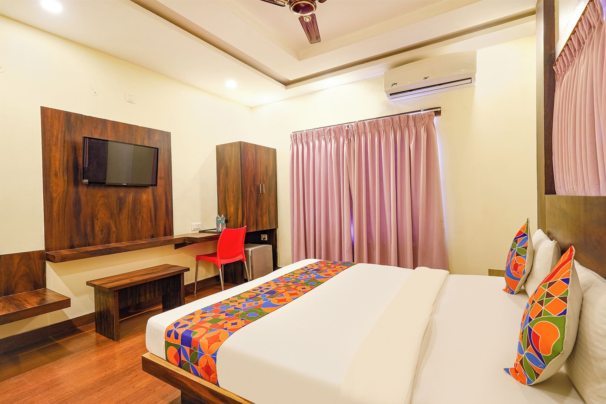 A hotel room with a bed, a desk with a chair, a TV mounted on the wall, and an air conditioning unit.