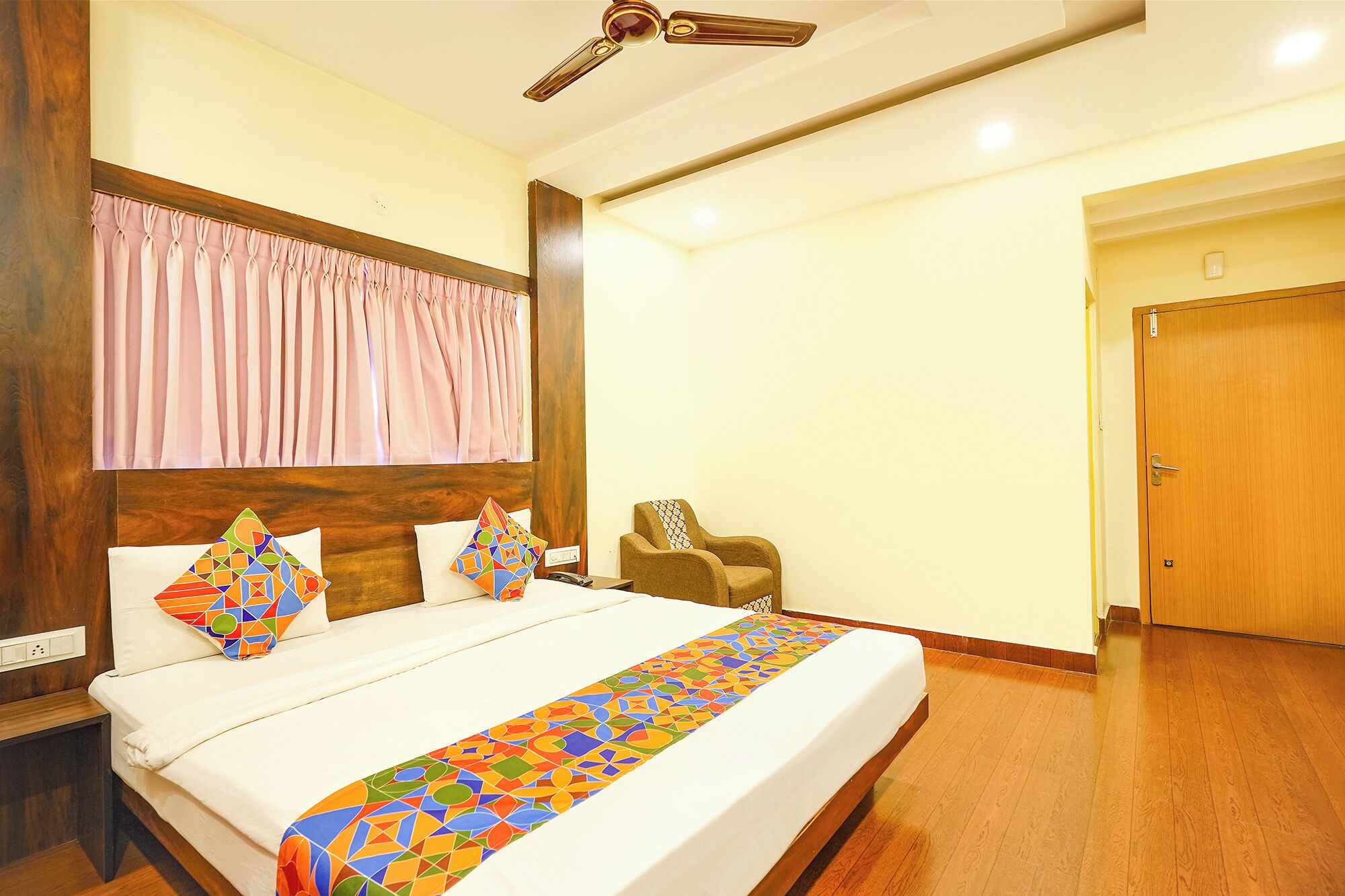 A hotel room with a bed, a wooden headboard, a ceiling fan, a chair, and a door.