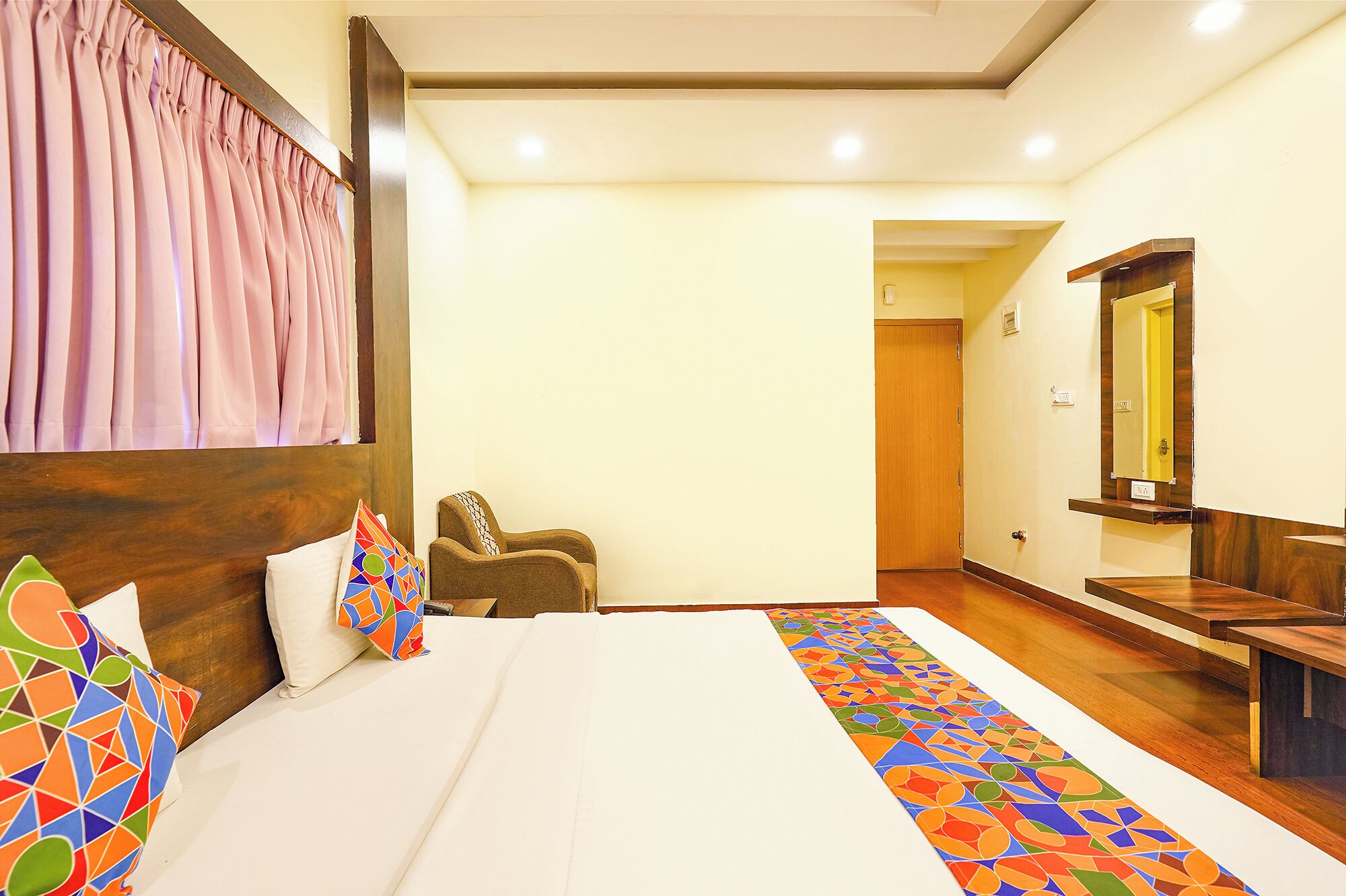 A hotel room with a bed, a chair, a wooden bench, a mirror, and a door.