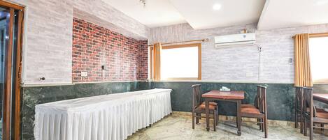 A room with a long table covered with a white cloth, two wooden chairs, a brick wall, and a window with curtains.