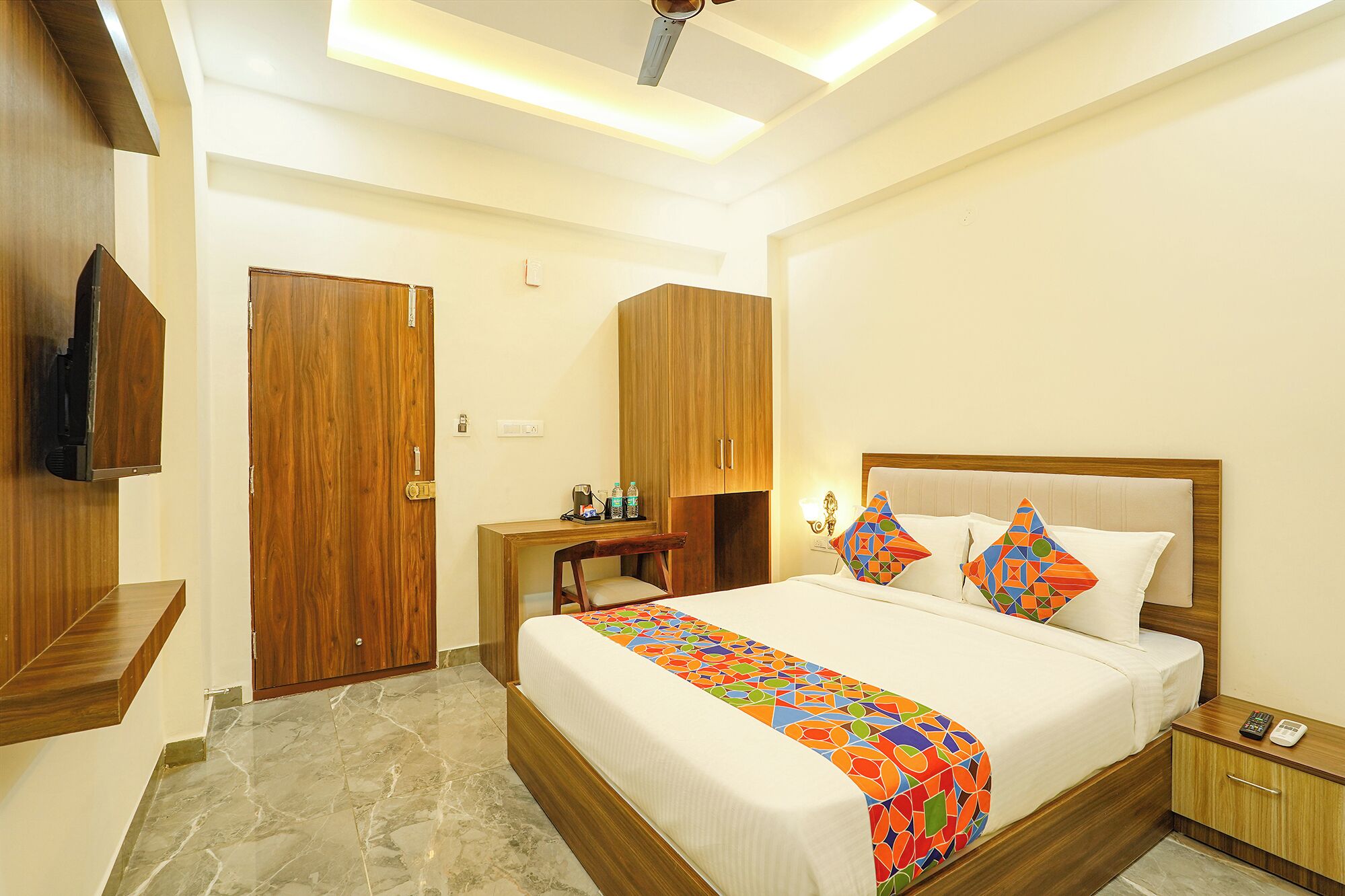 A hotel room with a bed, wooden wardrobe, a desk with a chair, a TV mounted on the wall, and a ceiling light fixture.