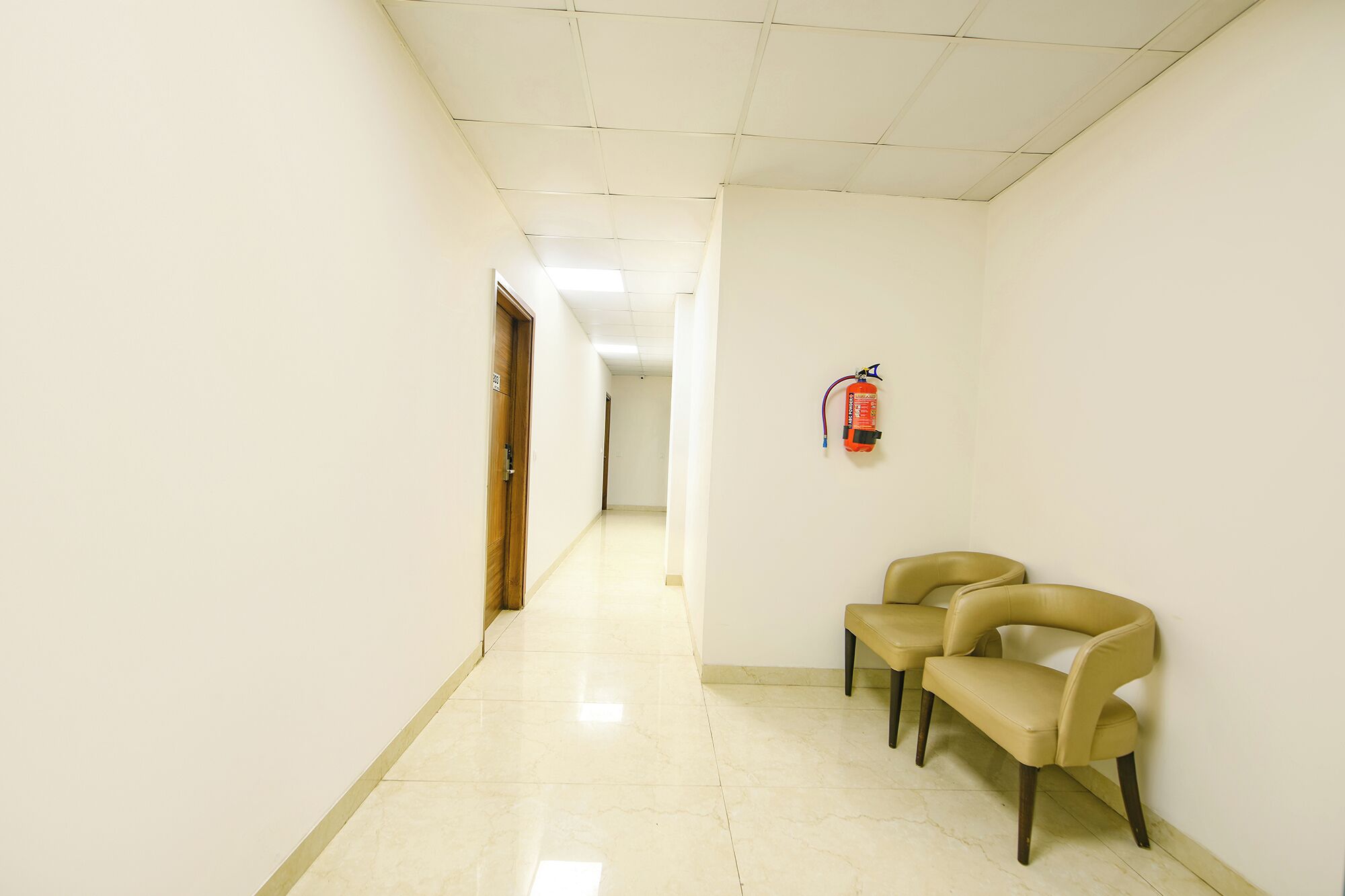 Um corredor com um extintor fixado na parede, duas cadeiras encostadas e uma porta no final.