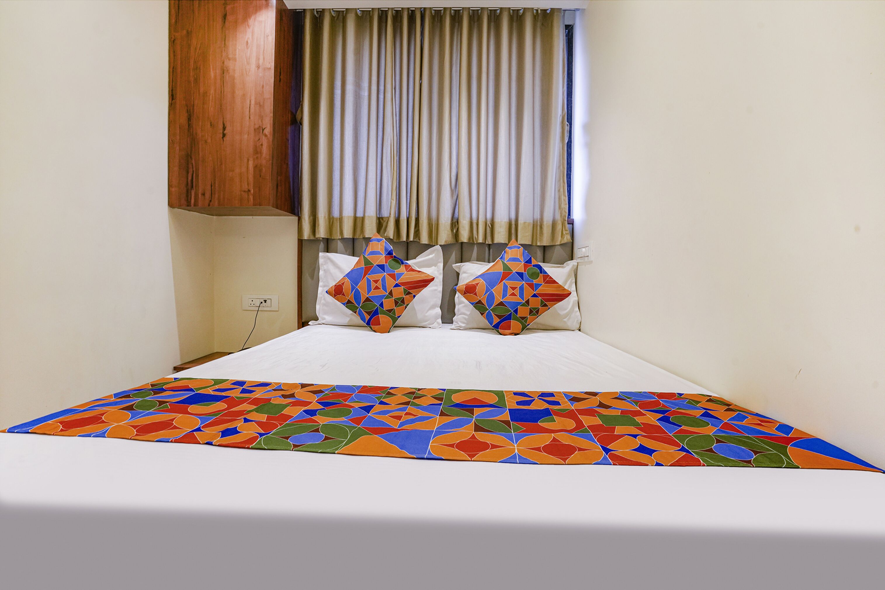 A bed with a colorful geometric-patterned bedspread, two decorative pillows, a wooden headboard, and a window with curtains.
