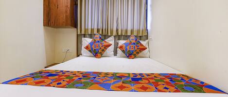 A bed with a colorful geometric-patterned bedspread, two decorative pillows, a wooden headboard, and a window with curtains.
