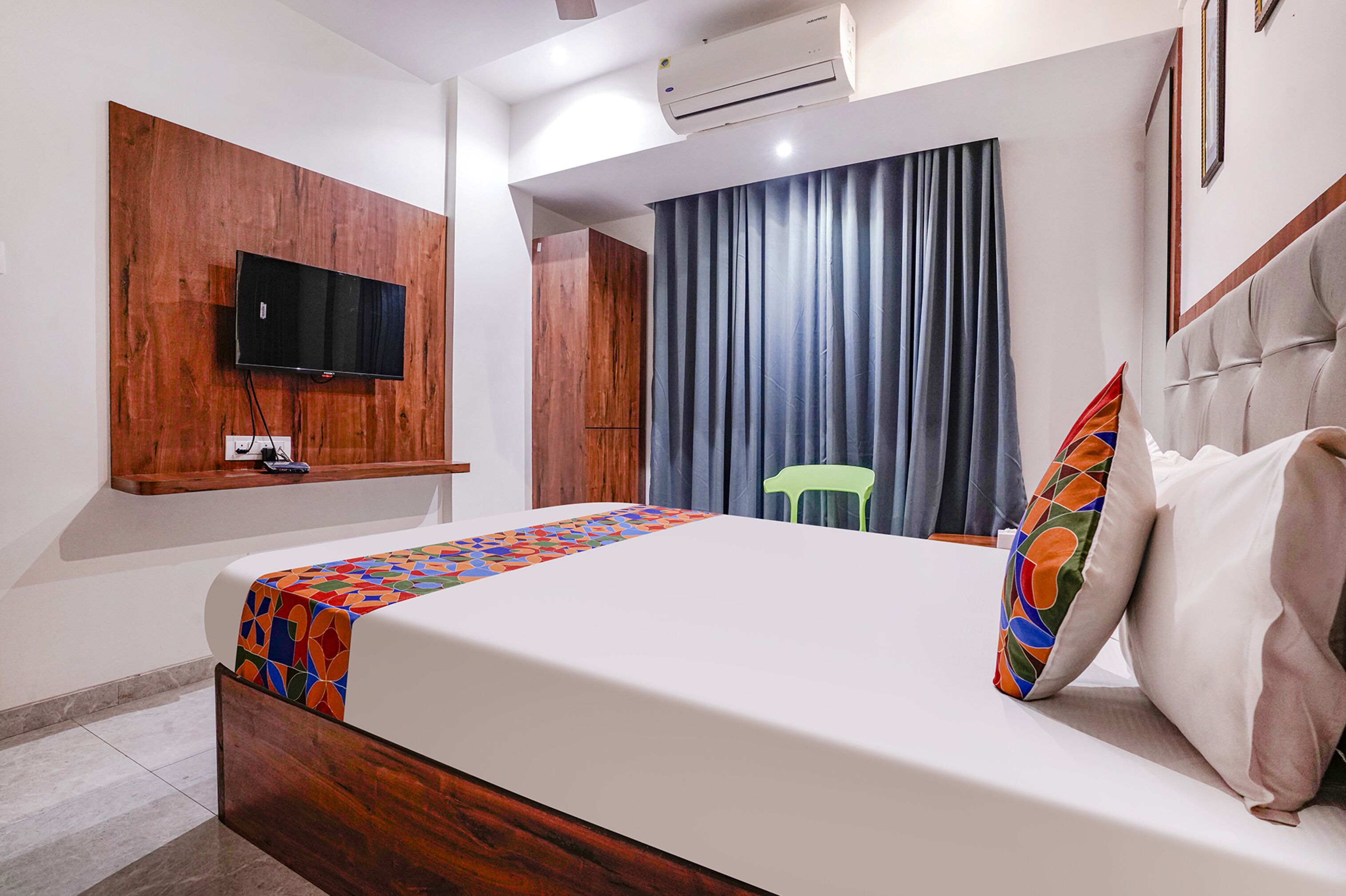 A hotel room with a bed, a mounted TV, a window with curtains, and an air conditioning unit.