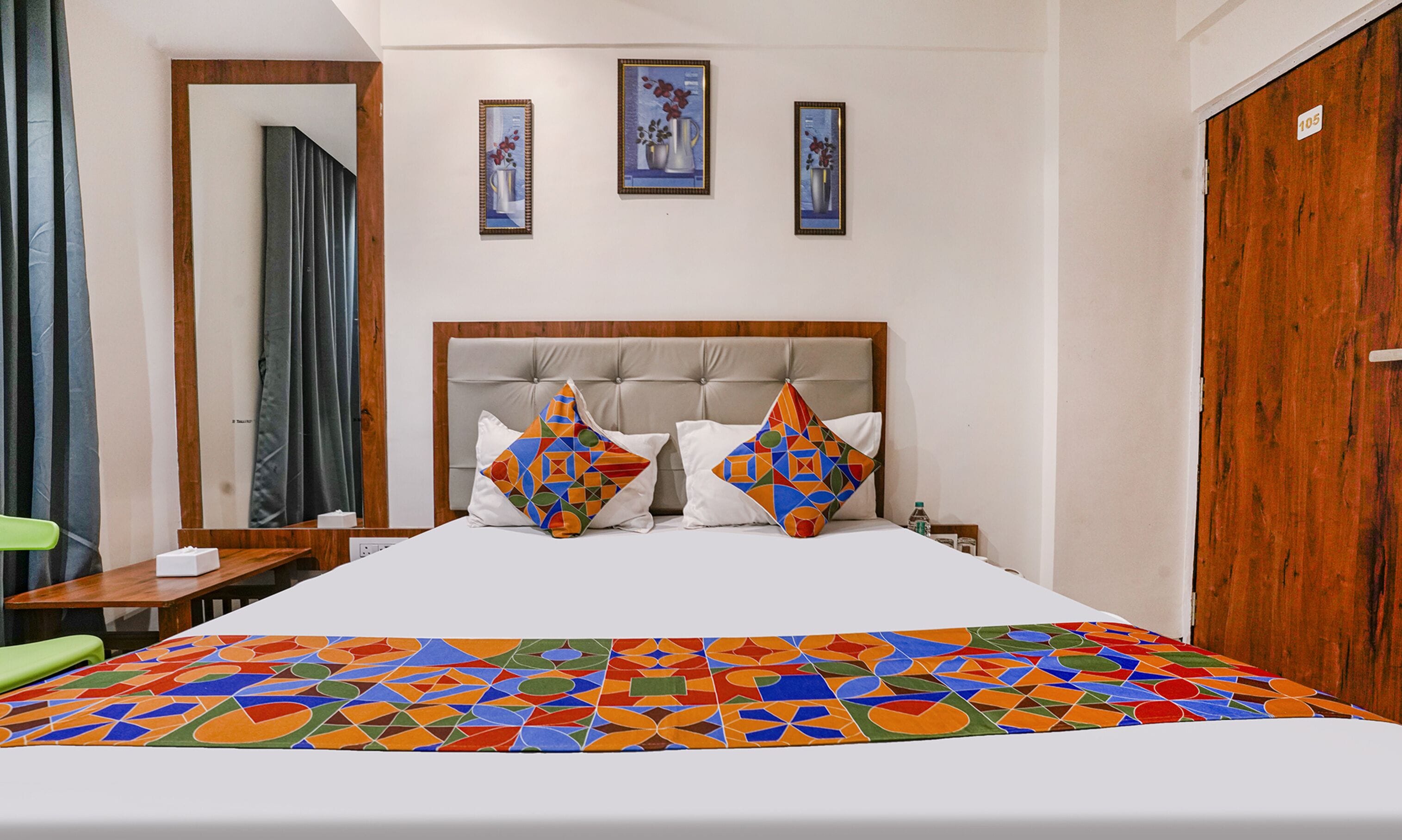 Habitación de hotel con una cama grande, cabecera de madera, dos almohadas coloridas, un armario de madera y tres cuadros en la pared.