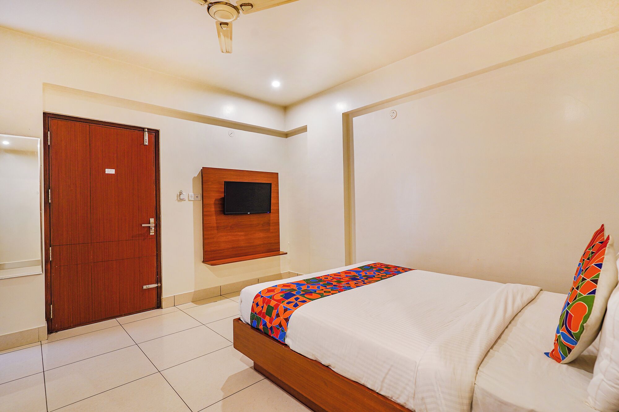 A bedroom with a bed, a wooden wardrobe, a television mounted on the wall, and a ceiling fan.