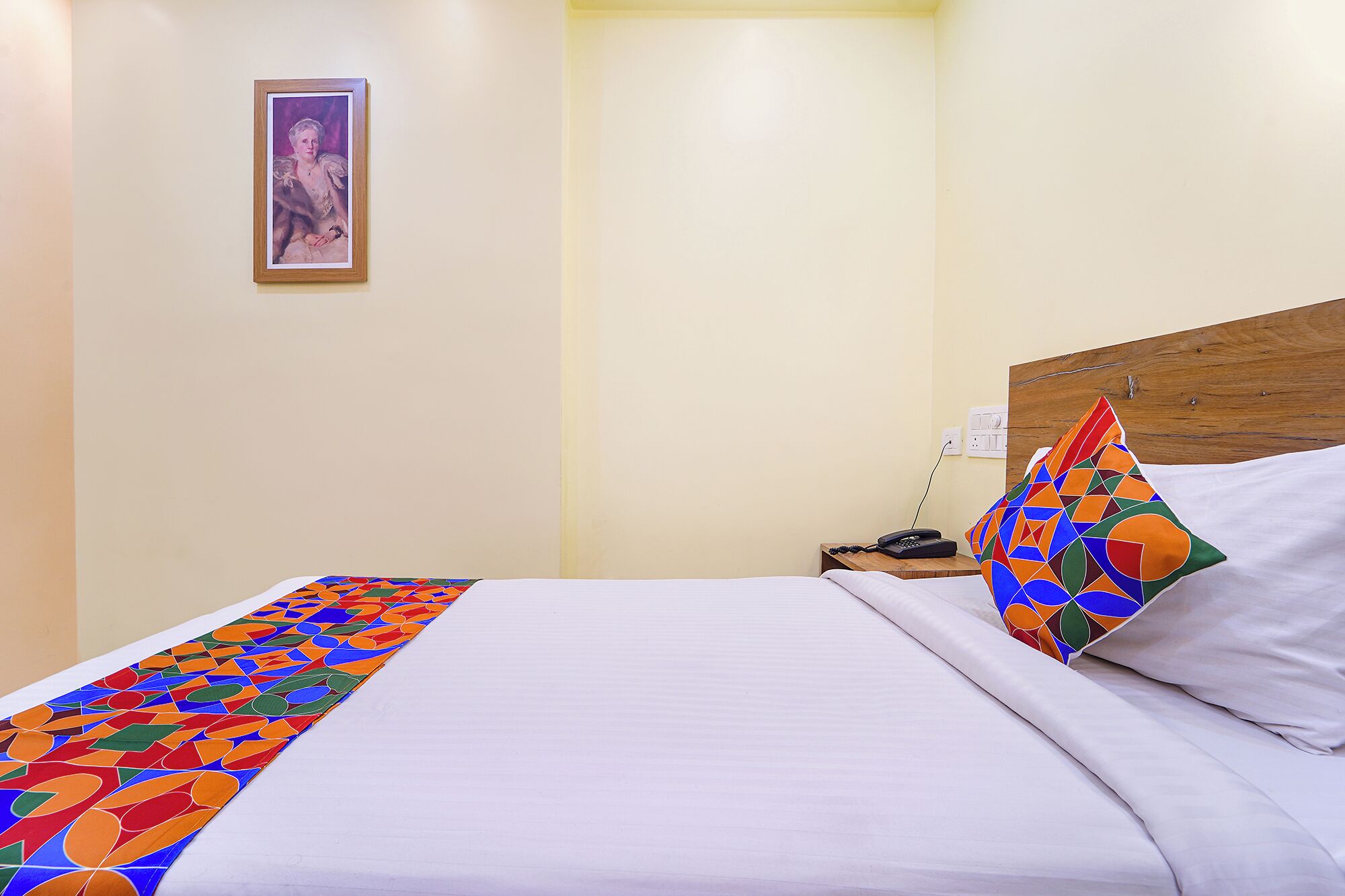 A neatly made bed with a colorful geometric-patterned bedspread, a wooden headboard, and a single framed picture on the wall.