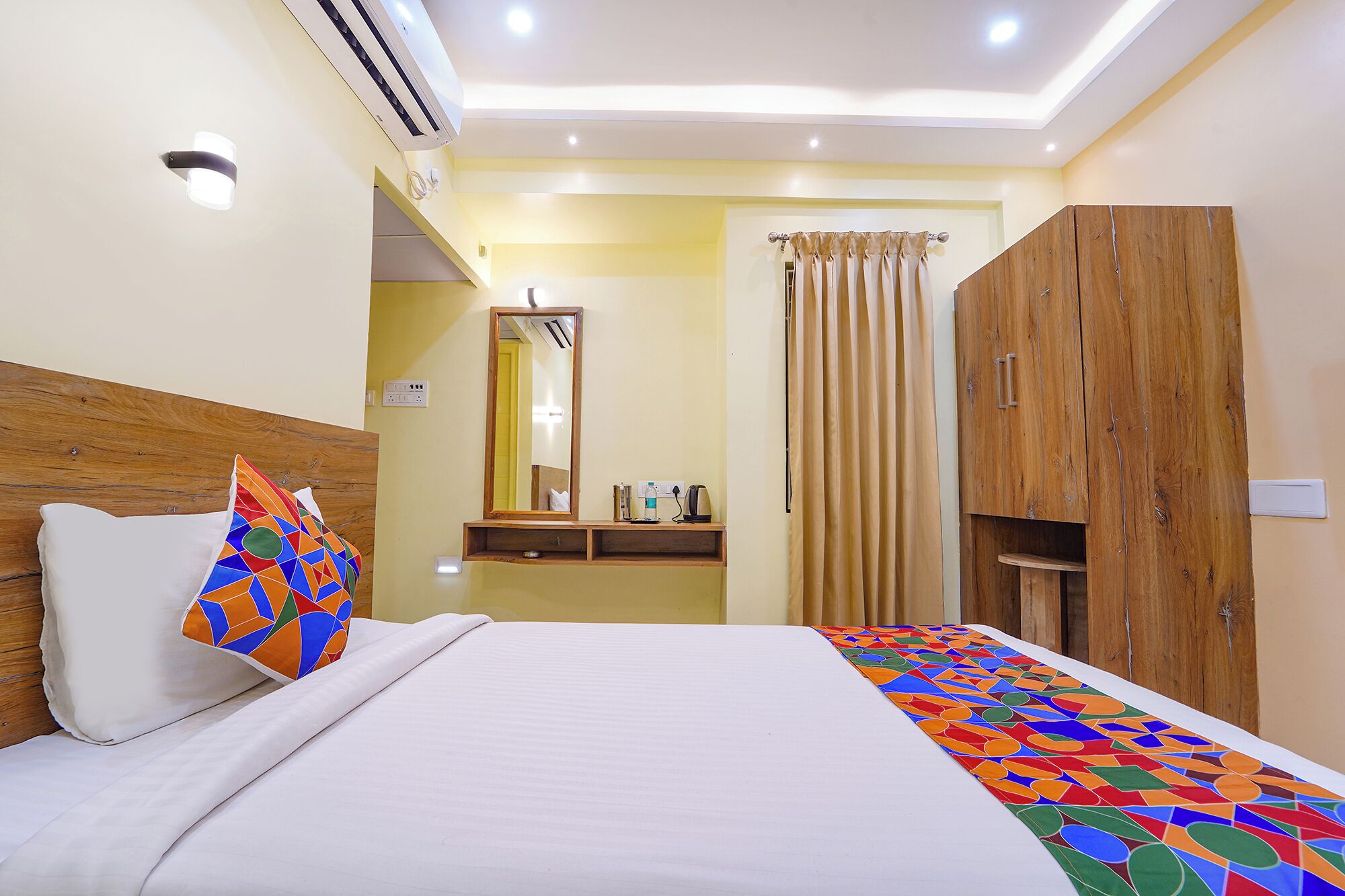 A hotel room with a bed, a wooden wardrobe, a mirror, a bedside table, and a window with curtains.