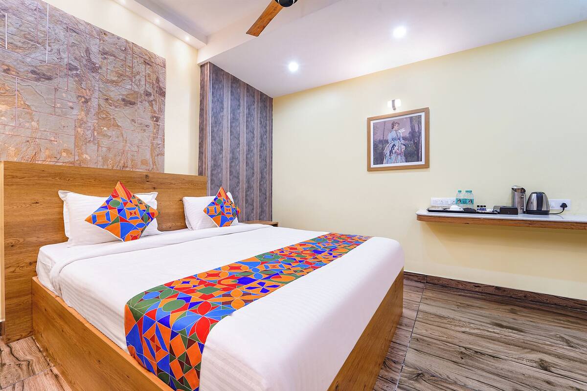 A hotel room with a wooden headboard, a bed with white bedding and colorful geometric-patterned throw, a wooden floor, a small shelf with a kettle and water bottles, and a framed picture on the wall.
