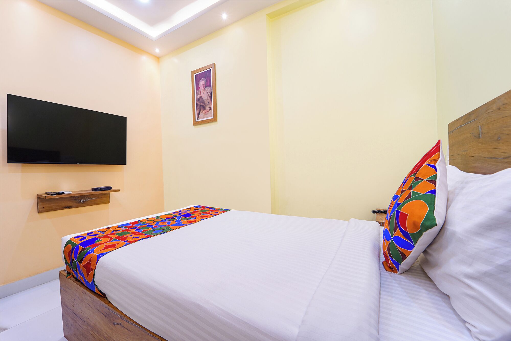 Habitación de hotel con una cama, un televisor montado en la pared, un pequeño estante de madera y un cuadro enmarcado.